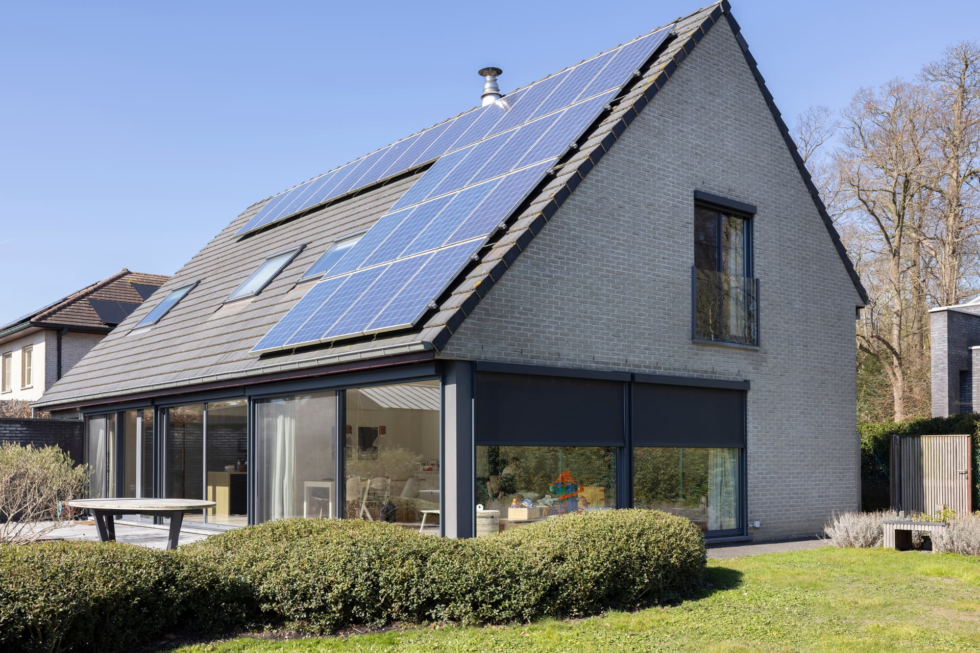 Maison moderne de plain-pied avec grandes baies vitrées coulissantes et stores extérieurs orange donnant sur un jardin verdoyant.