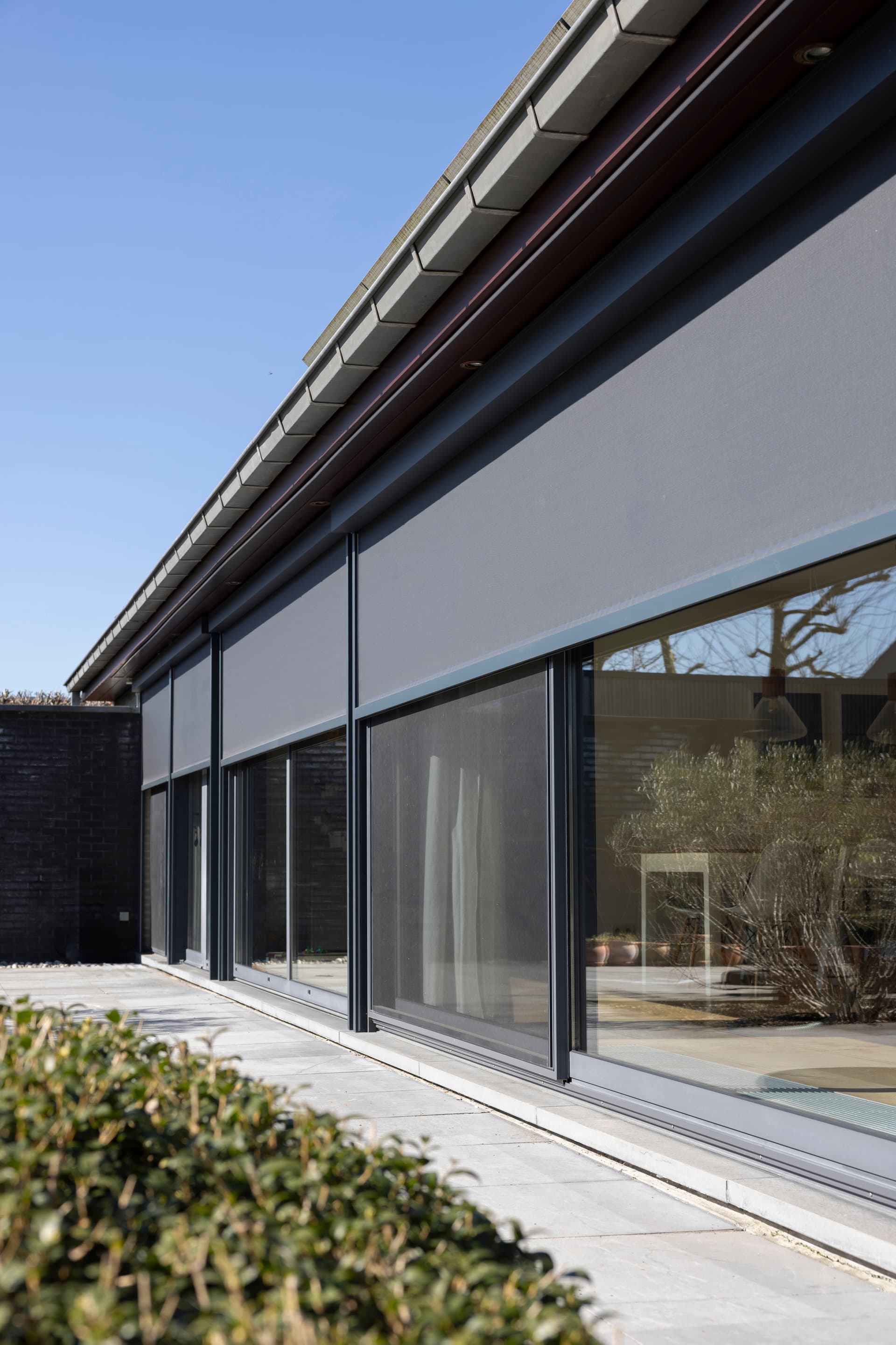 Maison moderne de plain-pied avec grandes baies vitrées coulissantes et stores extérieurs orange donnant sur un jardin verdoyant.