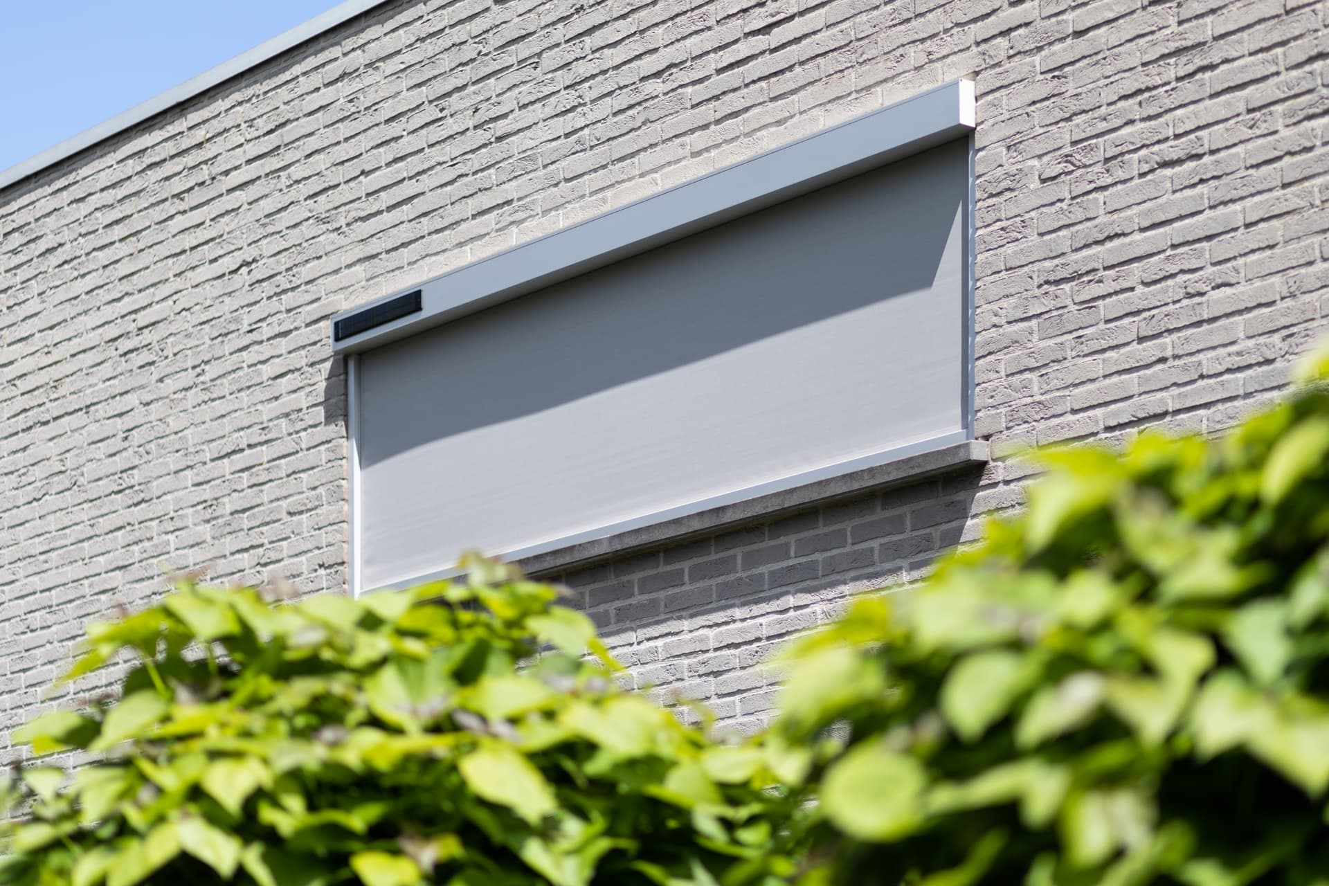 Brustor Solar‑ZIP‑screen met geïntegreerd zonnepaneel aan een moderne bakstenen gevel voor autonome zonwering