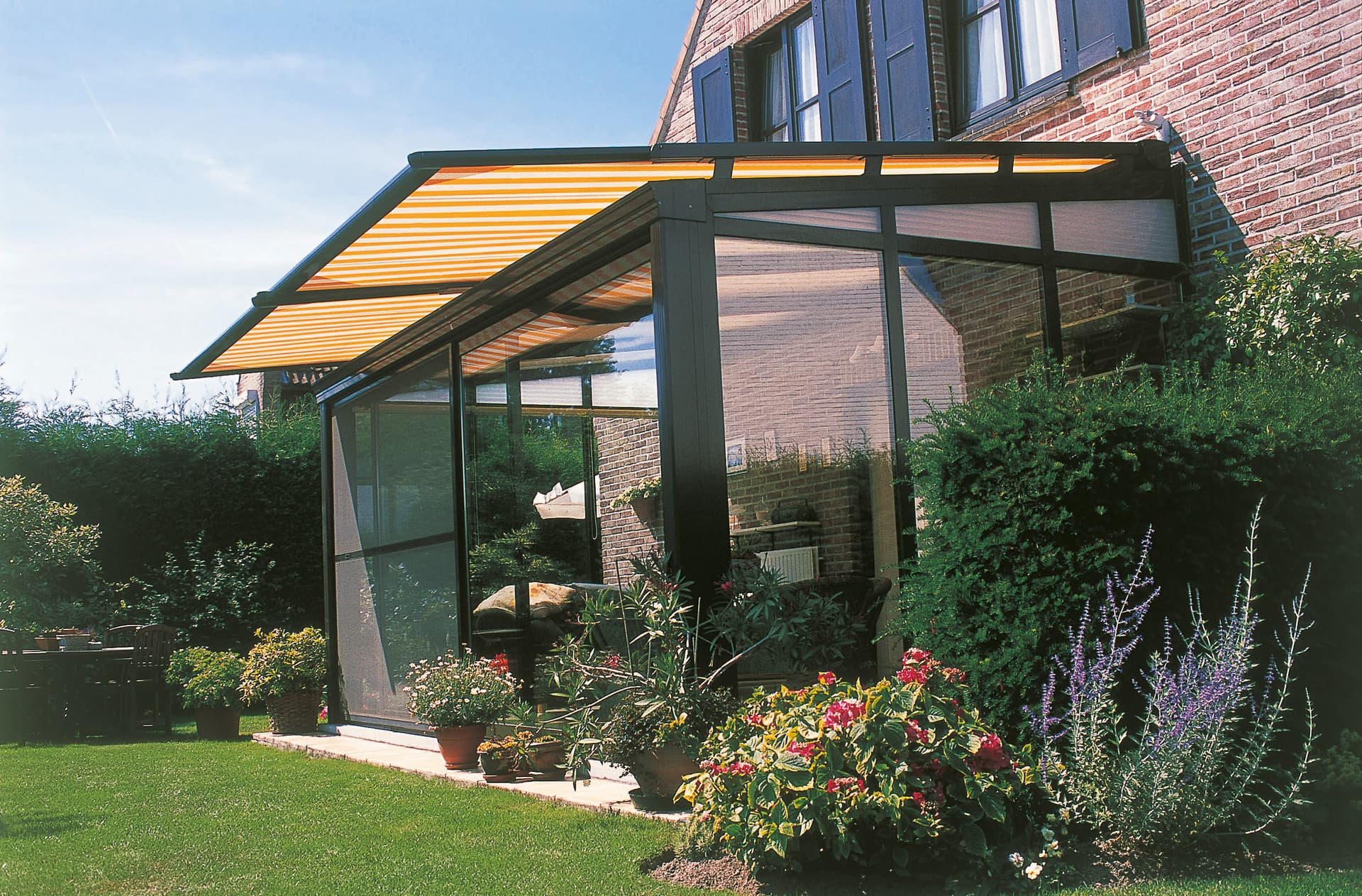 Brustor B126 conservatory awning installed above an irregularly shaped glass veranda, providing exterior sun protection in a lush garden setting
